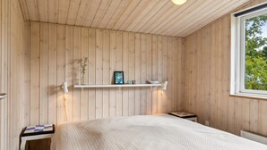 Een kamer met een houten lambrisering, een bed met een witte quilt, een klein tafeltje met een quilt, een plank met decoratieve objecten, en een raam met uitzicht op bomen.