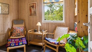 Een gezellige kamer met houten wanden, een groot raam met uitzicht op groen, een houten bed, een stoel met een kleurrijk kussen en een klein tafeltje met een lamp.