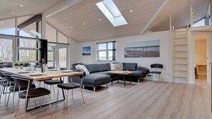 A spacious living area with a wooden floor, a dining table with chairs, a grey sofa, and a large window with a view of the beach.