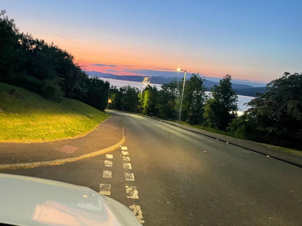Una carretera con una curva, rodeada de vegetación y un atardecer al fondo.