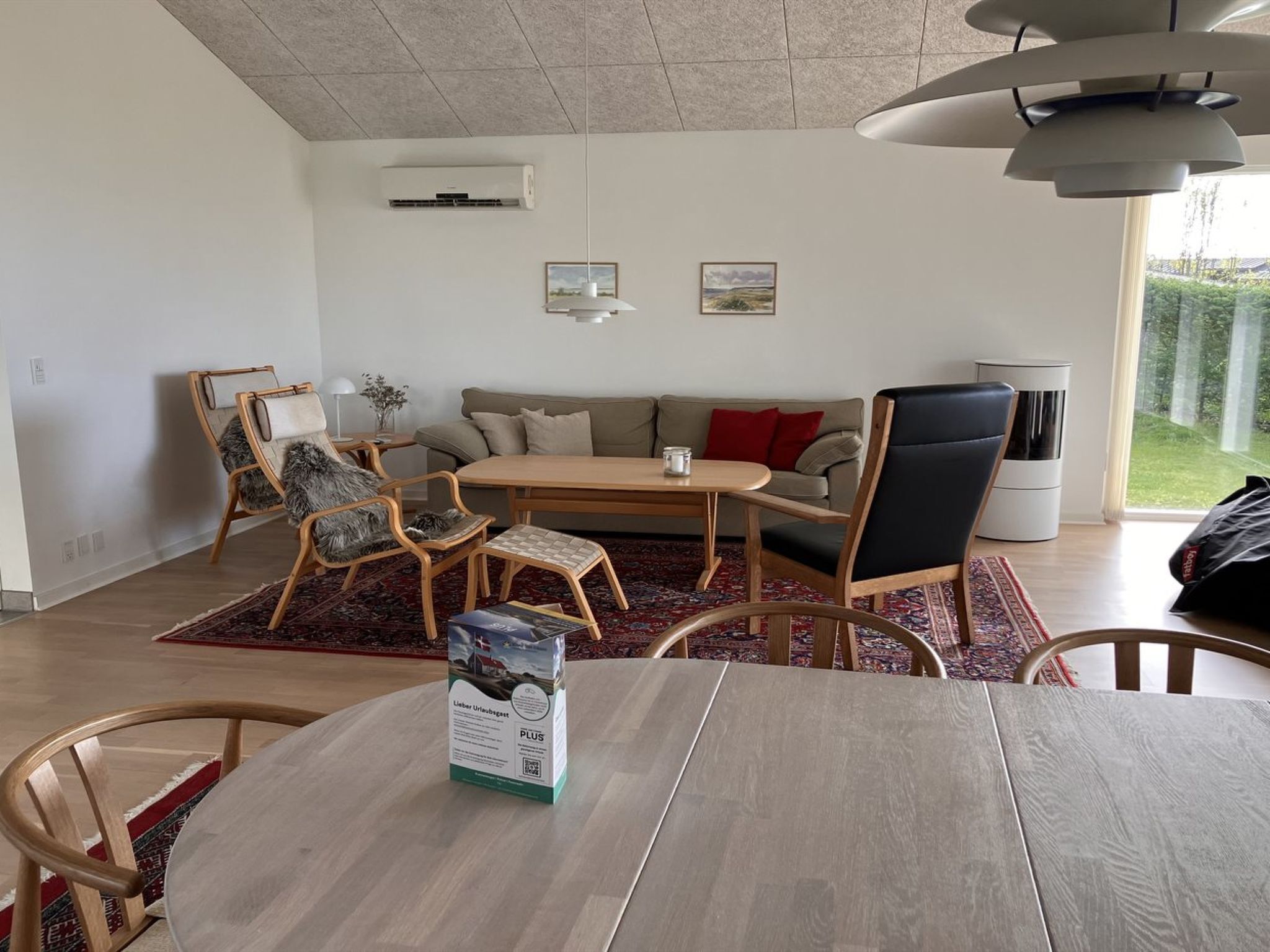 Ett modernt vardagsrum med ett matbord i trä, två soffor och ett soffbord. Det finns två stolar, en matta och ett fönster med utsikt över grönområden.