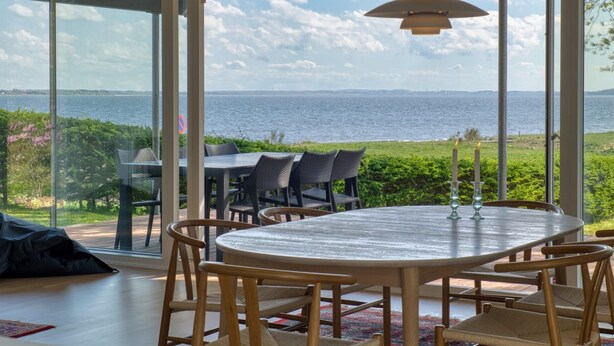 Comedor con mesa redonda y sillas, ventanas amplias con vista al mar y vegetación.