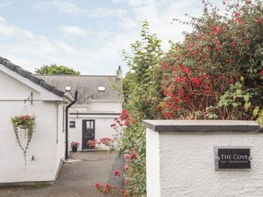 Un bâtiment blanc avec une porte noire, une enseigne indiquant « The Cove Flat 1, 1 Treadmore Road », et un jardin avec des fleurs rouges.