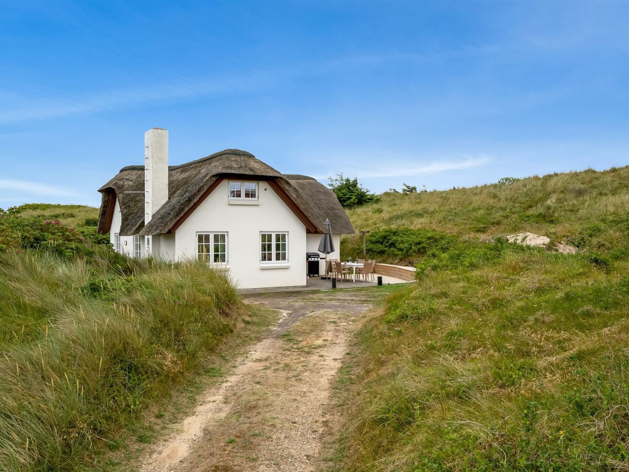 Et hvidt hus med stråtækt tag, en grussti der fører til huset, og en græsdækket bakke i baggrunden.