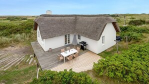 Une maison à toit de chaume avec une terrasse en bois et une table ainsi que des chaises à l’extérieur.