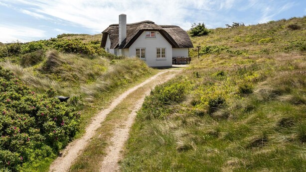 Une maison blanche avec un toit de chaume, située sur une colline verdoyante et accessible par un sentier de terre.