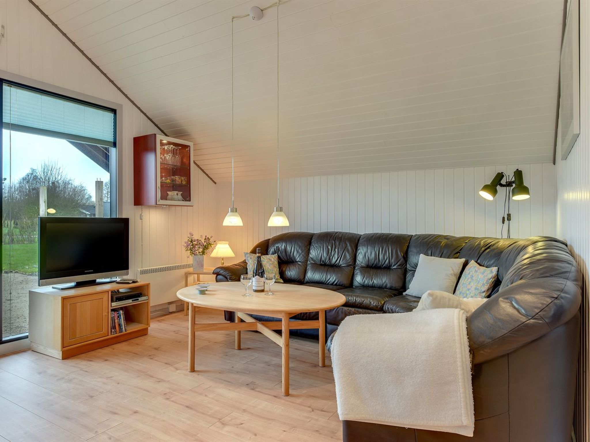 A modern living room with a leather sofa, a flat-screen TV, wooden flooring, and pendant lights.