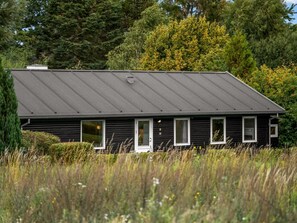 Een zwart houten huis met schuin dak, omringd door hoog gras en bomen.
