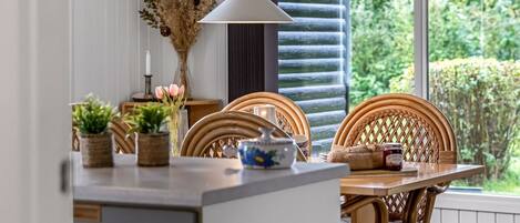 A dining area with a table set for two, featuring wicker chairs, a vase with dried flowers, and a teapot.