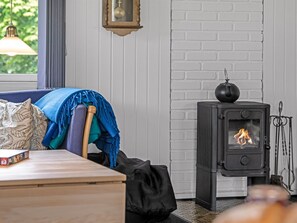 A cozy room with a wooden table, a chair with a blanket, a black wood-burning stove, and a window with a hanging light.