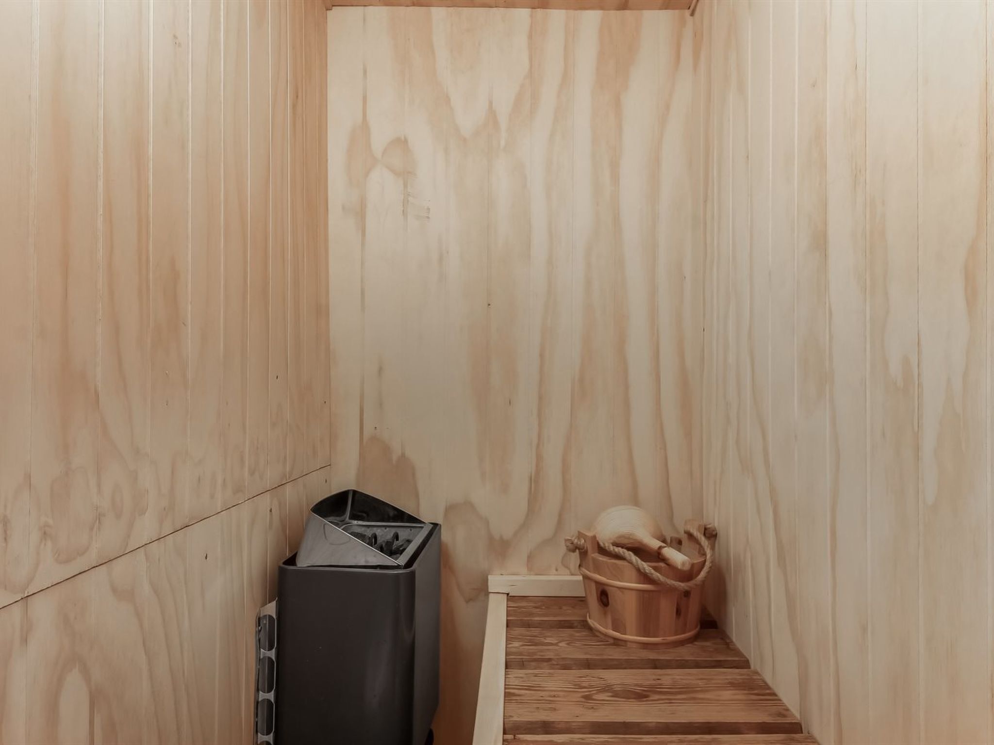 Il s’agit d’une petite pièce fermée, aux murs en bois et au plafond en pente. Une poubelle noire est placée contre le mur gauche, et un seau en bois muni d’une anse en corde se trouve sur le sol.