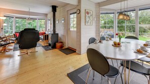 Une pièce chaleureuse avec un plancher en bois, une table à manger avec des chaises, un fauteuil noir, un canapé avec un coussin rouge, une cheminée et de grandes fenêtres donnant sur un paysage verdoyant.