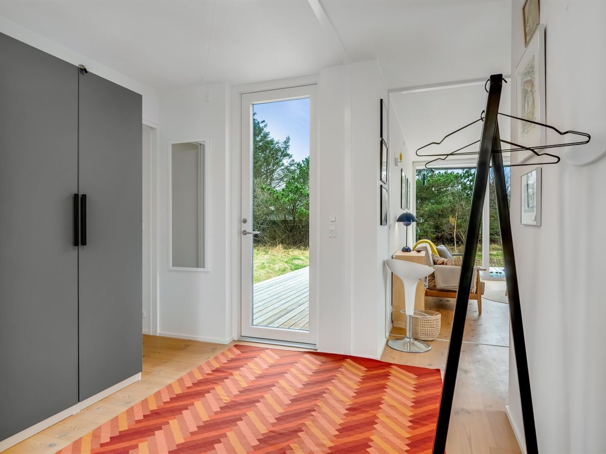 A modern interior with a grey wardrobe, a herringbone-patterned rug, a hanging coat rack, and a view of an outdoor deck.