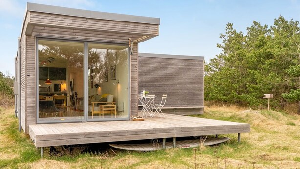 Une cabane moderne dotée d’une entrée avec porte en verre, d’une terrasse en bois et d’un espace repas aménagé avec du mobilier d’extérieur.