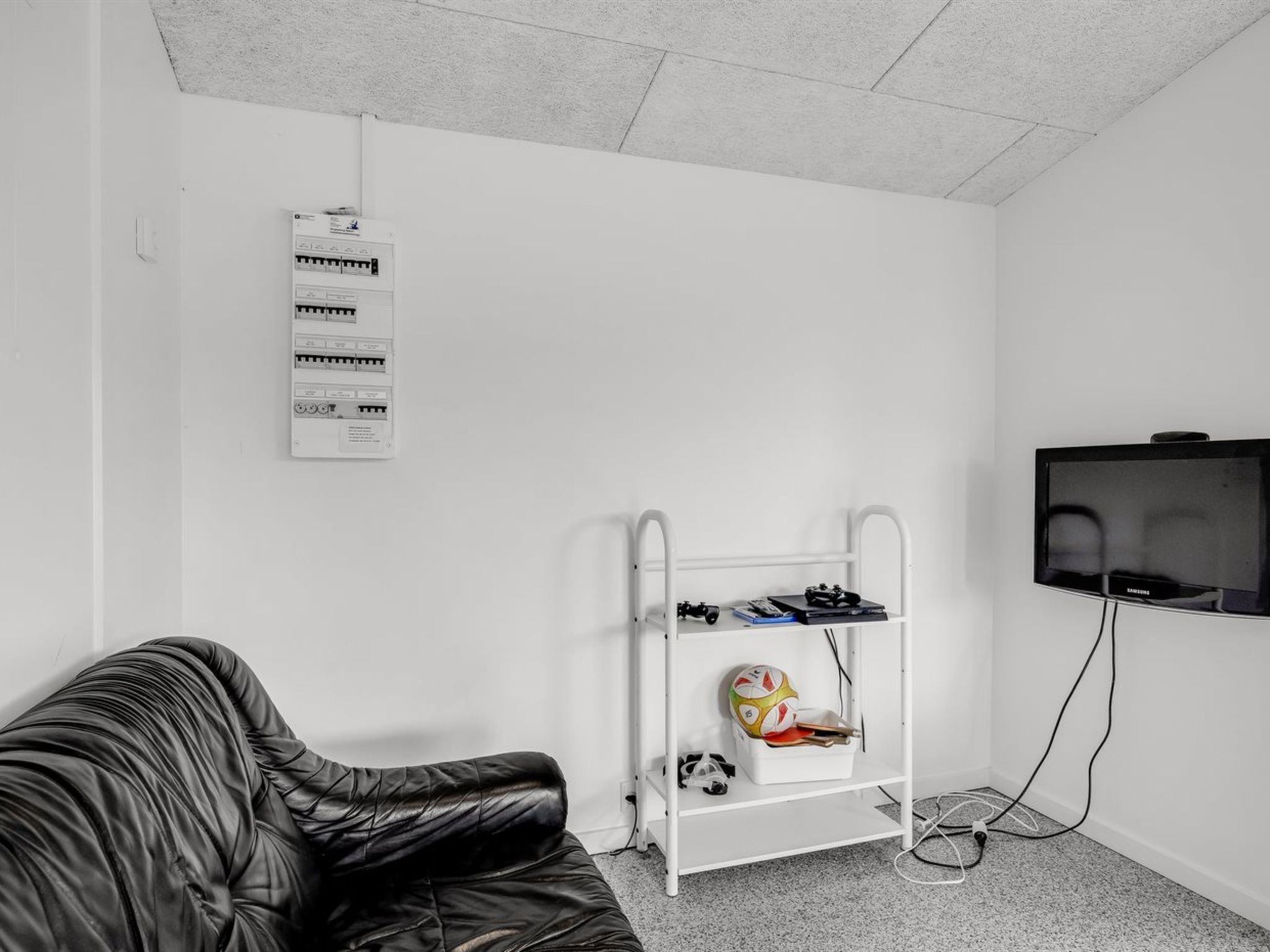 Ett rum med en vit vägg, en vit hylla med sportutrustning, en svart lädersoffa och en tv på väggen.
