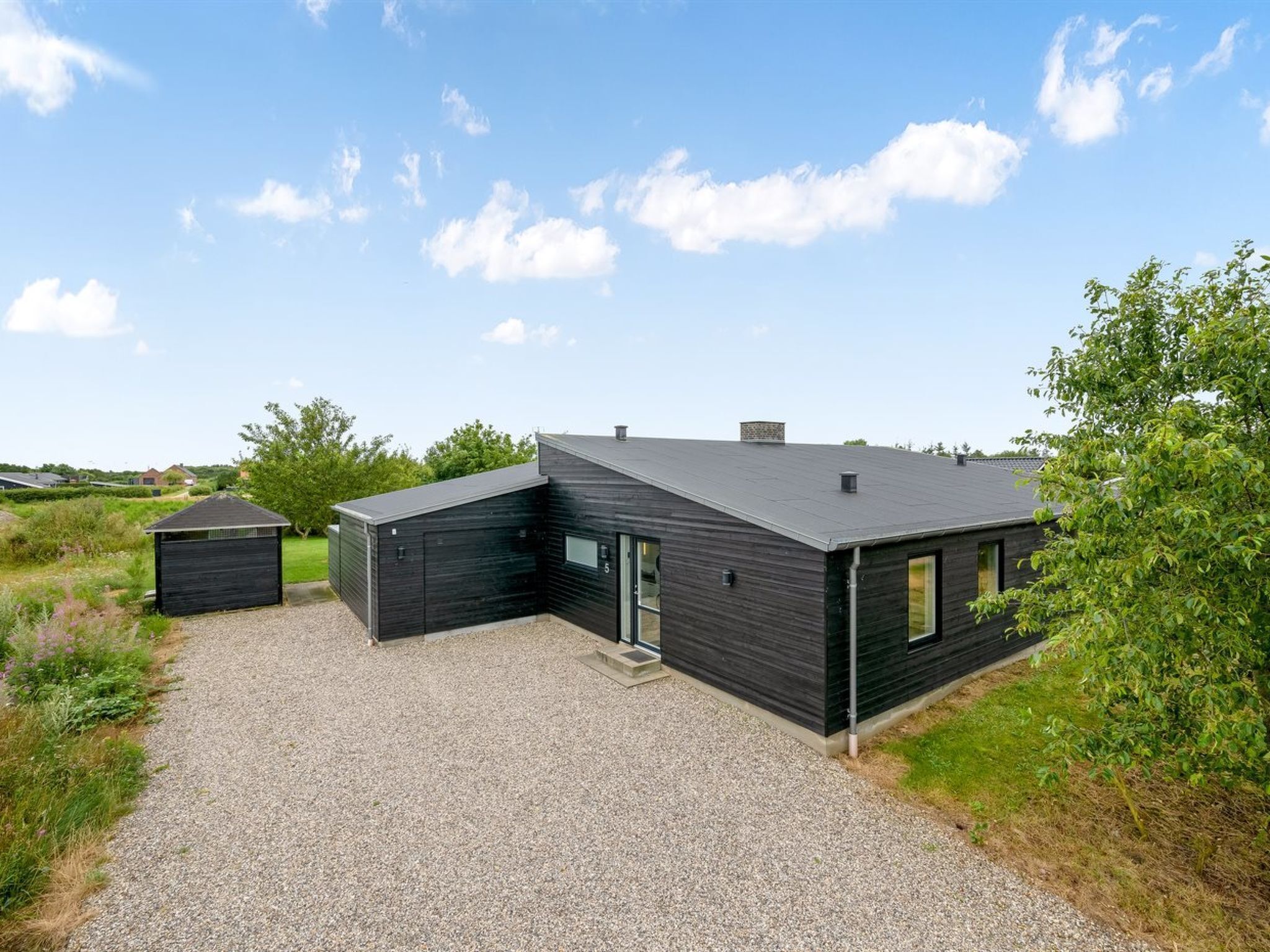 En moderne, sort bygning med grusvej, et lille skur og en græsplæne med træer.