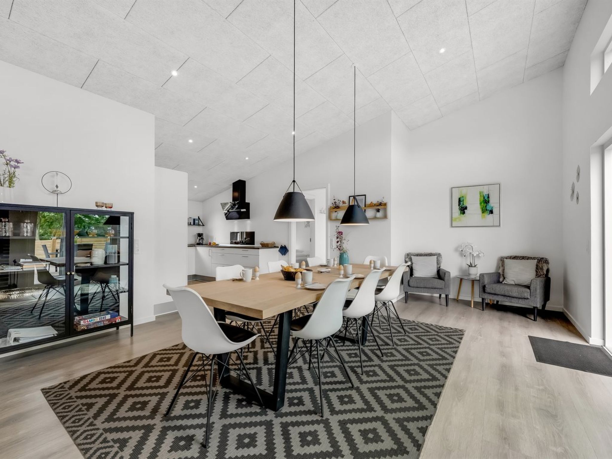 A modern kitchen with a dining area, featuring a large wooden table, white chairs, and black pendant lights.