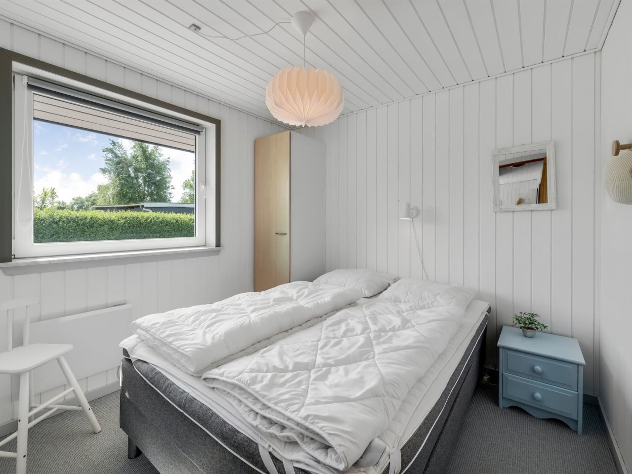 A bedroom with a bed, a desk, a dresser, and a window with a view of greenery.