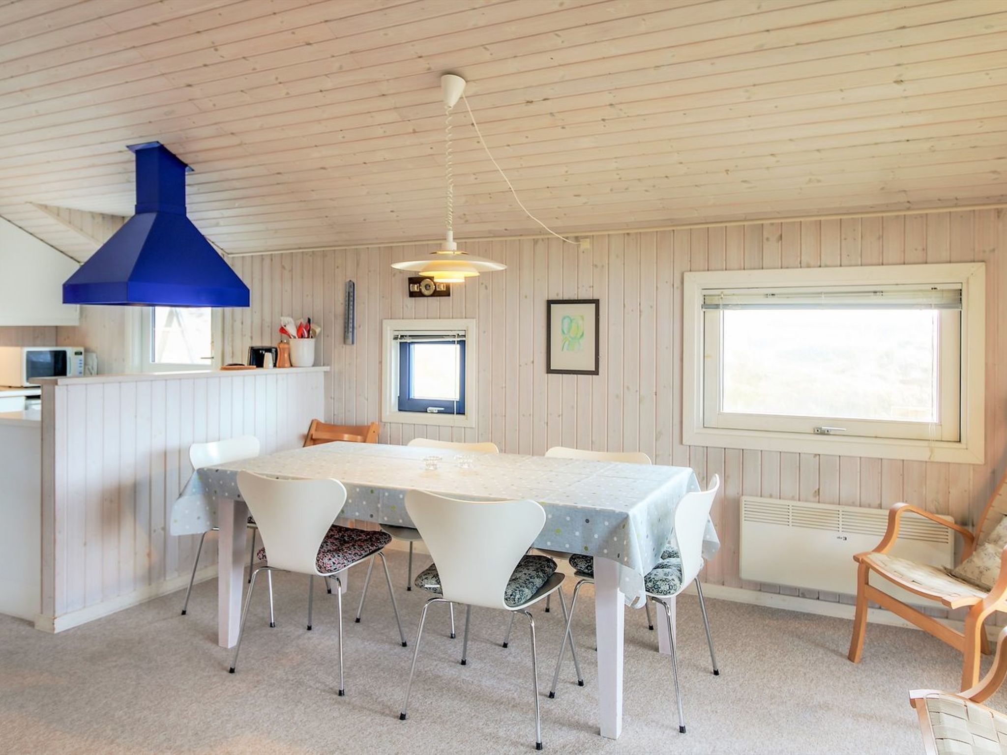Un coin repas avec une grande table, des chaises blanches, une bouche d’aération bleue au plafond et une fenêtre avec des stores.