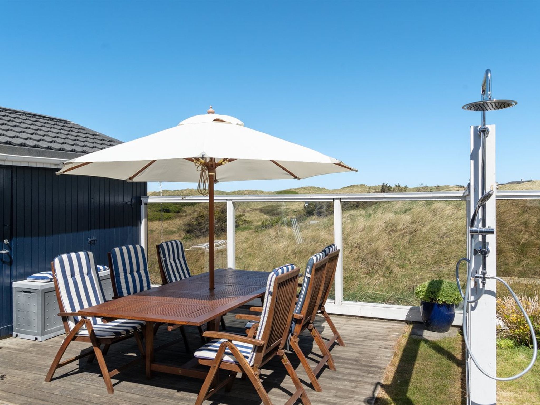 A deck with a wooden table and striped chairs, a white umbrella, and a shower.