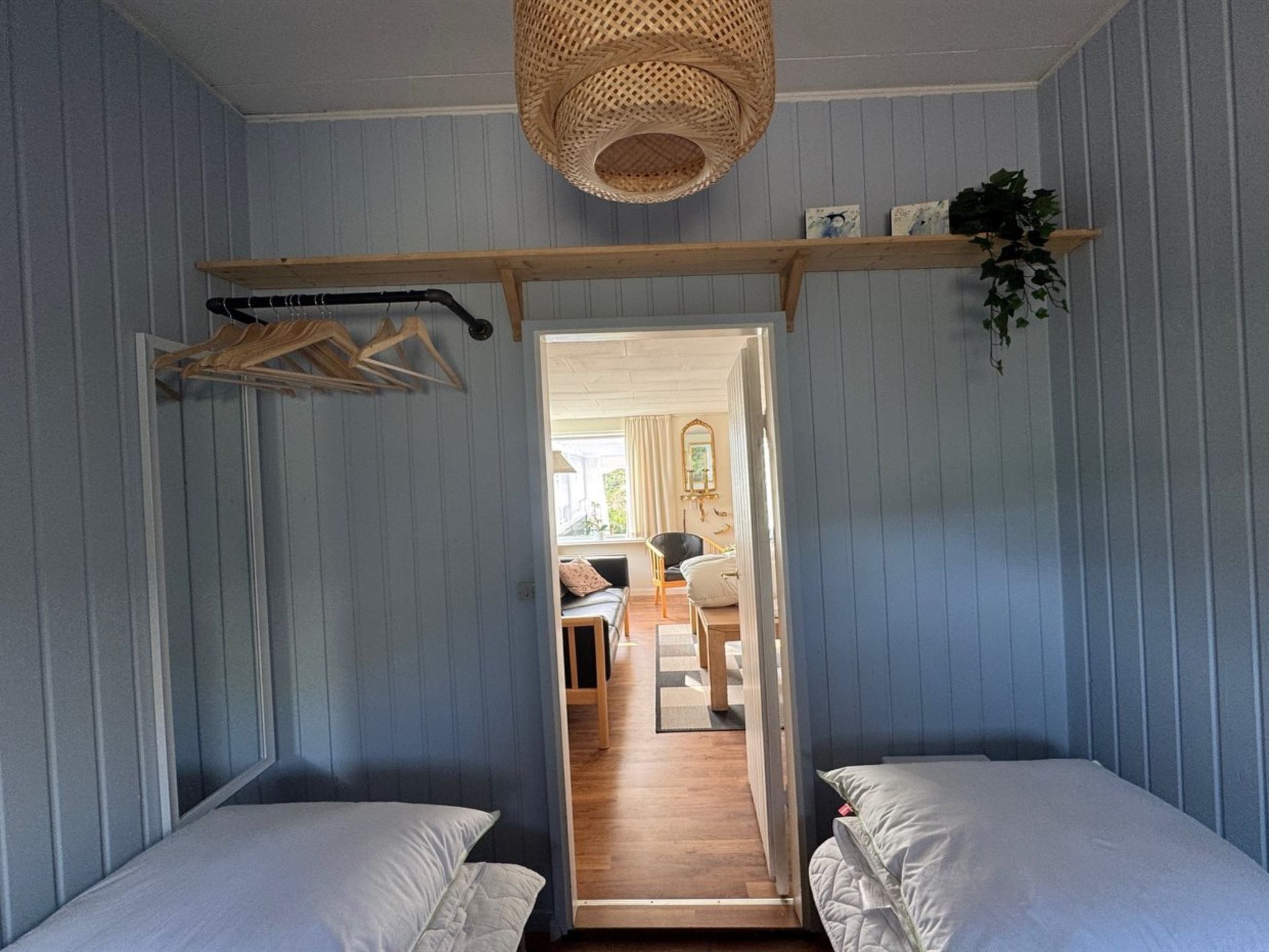 Ein Schlafzimmer mit zwei Betten, einem hölzernen Regal mit einer hängenden Pflanze und Blick in einen angrenzenden Wohnbereich.
