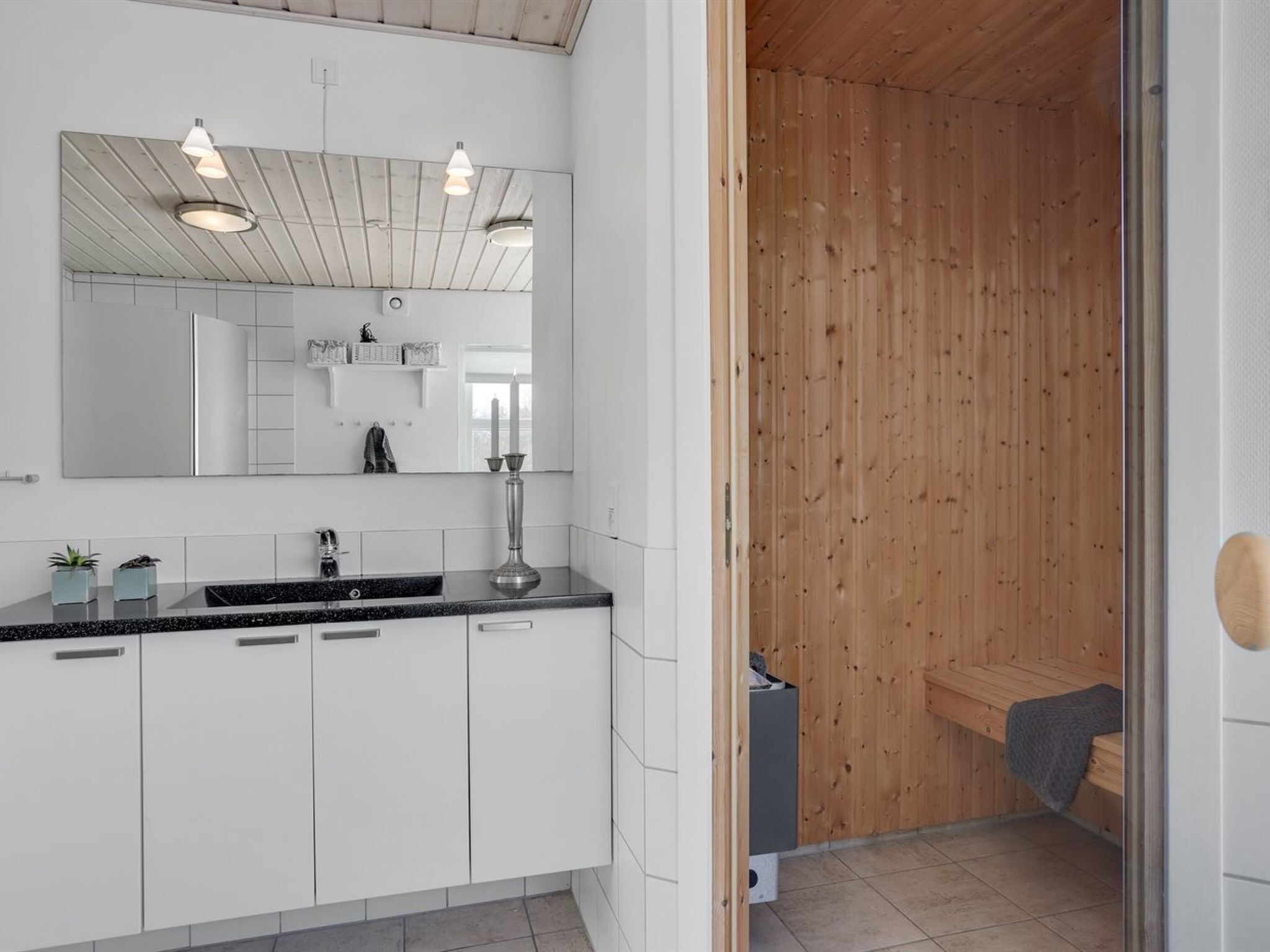 A modern bathroom with a large mirror, white cabinets, a black countertop, and a wooden sauna area.
