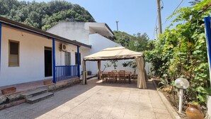 A covered patio with a table and chairs, a white building with blue railings, and a hillside in the background. - Room with breakfast included in cottage Holiday home in Palmi Pietrenere. (Palmi)