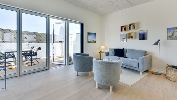 Een ruime woonkamer met een grote schuifpui naar een terras buiten, een grijze bank, twee fauteuils en een vloerlamp.