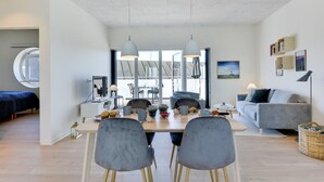 Een moderne woonkamer met een eethoek, met een rond raam, grijze gestoffeerde stoelen, een grijze bank en een groot raam dat natuurlijk licht binnenlaat.