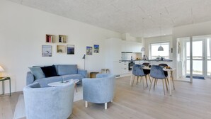 Een moderne woonkamer met een bank, twee fauteuils, een eethoek met tafel en stoelen, een keuken op de achtergrond, en een groot raam dat natuurlijk licht binnenlaat.