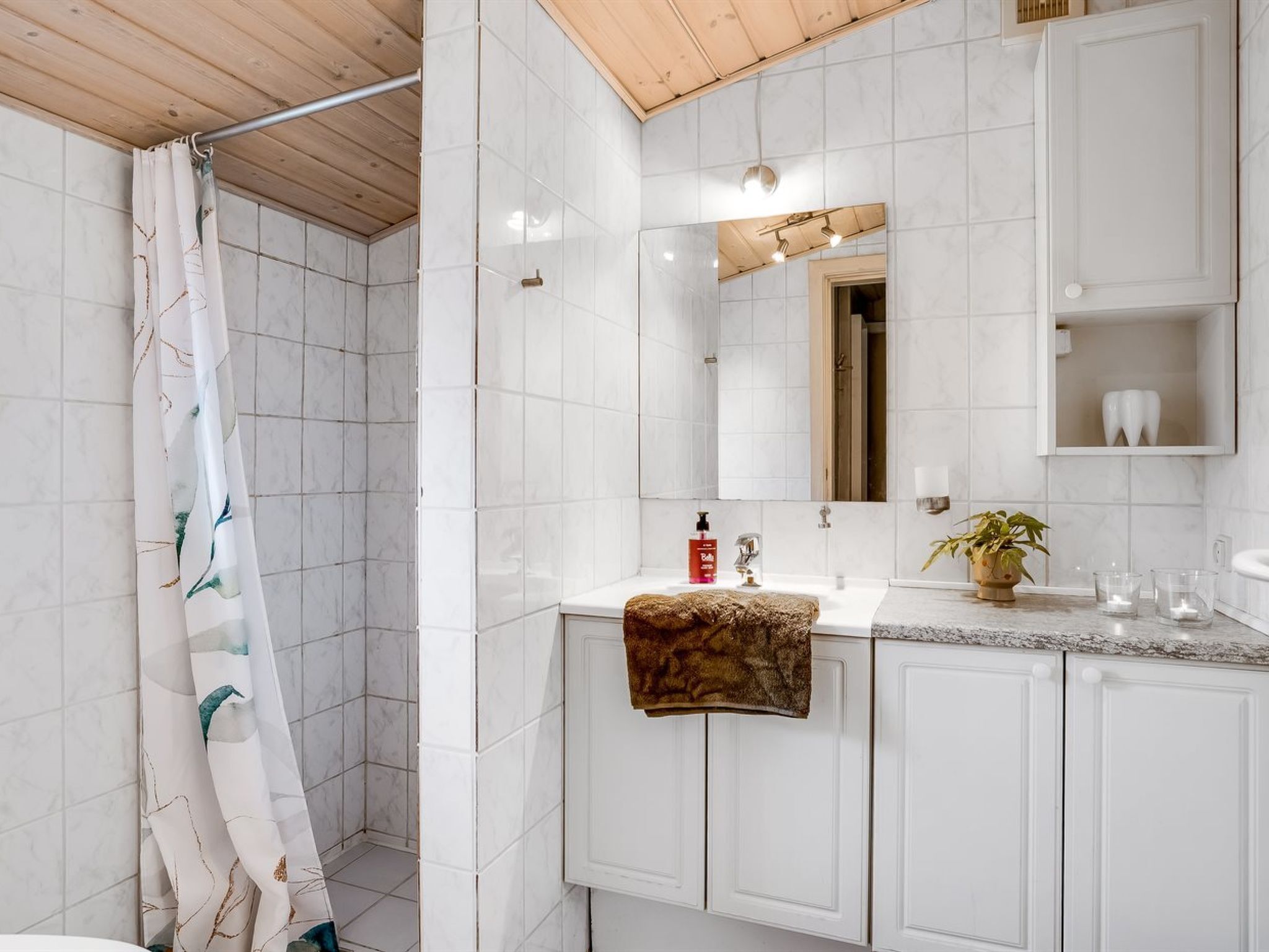Un baño con azulejos blancos, techo de madera, cortina de ducha, lavamanos con una toalla, espejo y una planta.