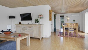 Un vaste salon doté d’un plafond en bois, d’un téléviseur à écran plat, d’un coin repas avec une table et des chaises en bois, et d’une cuisine ouverte sur le salon par une porte.