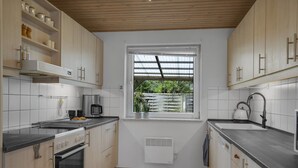 Une cuisine moderne avec des meubles de couleur bois clair, un four encastré, une machine à café et une fenêtre donnant sur un paysage verdoyant.