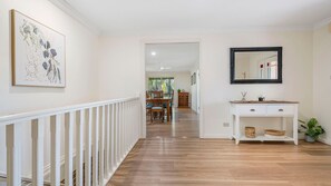 A hallway with a white railing, a wooden floor, a dining area with chairs and a table, a white console table with baskets, and a framed mirror on the wall.