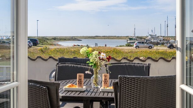 A table with wicker chairs, a vase with flowers, and two coffee cups with the text "IS KAF FE" on a terrace overlooking a beach with boats and cars. - "Idunn" - 100m to the fjord by Interhome (Vestervig)
