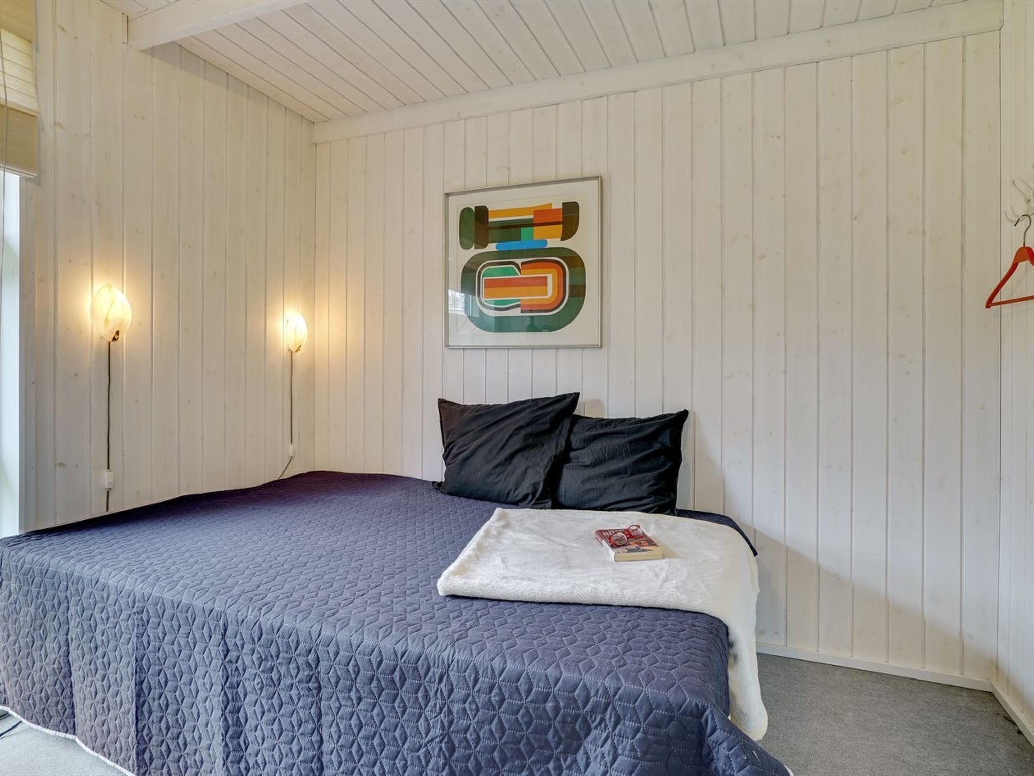 A bedroom with a bed, two wall-mounted lamps, a framed abstract painting, and a red hanger on the wall.
