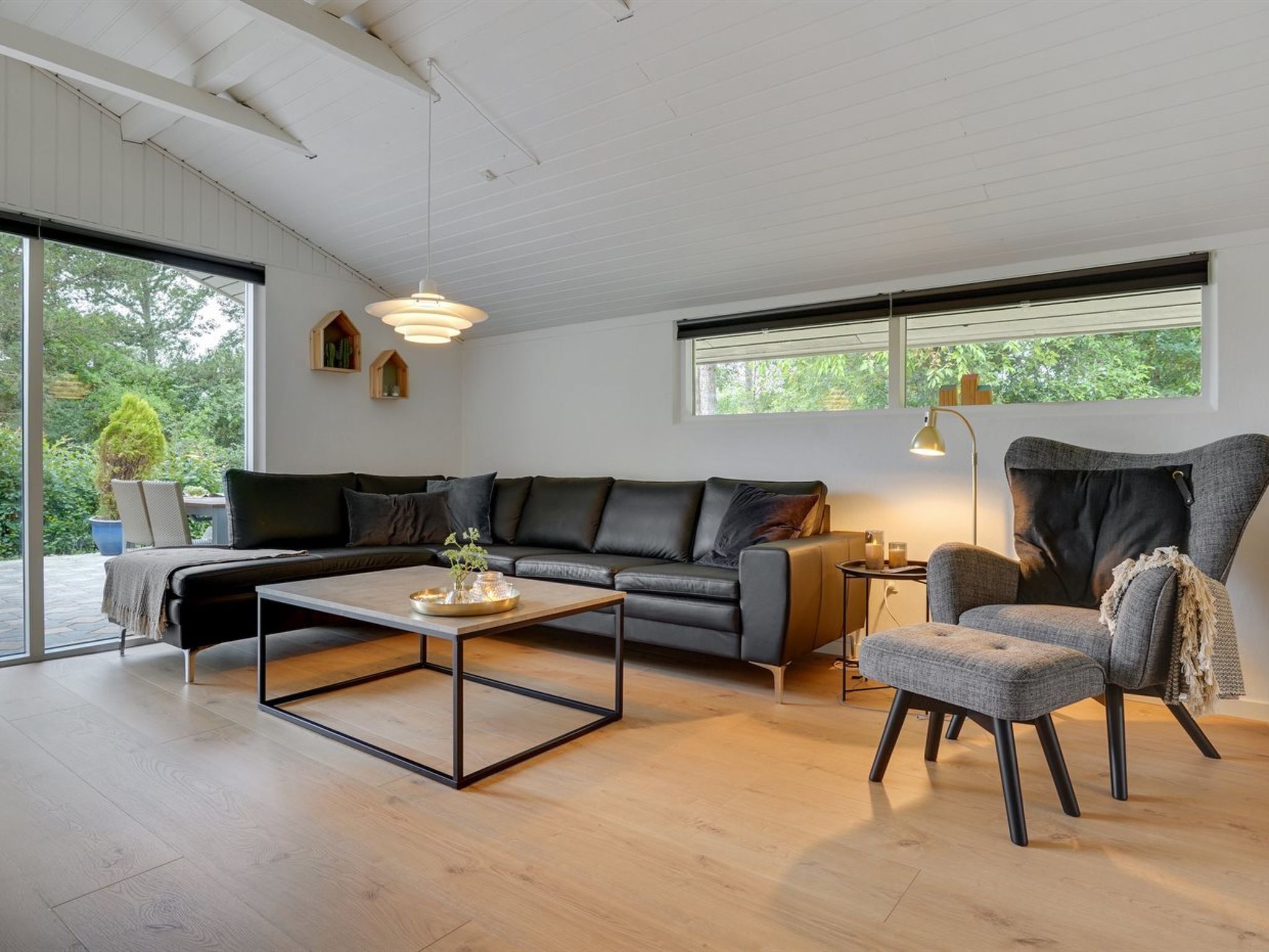 A modern living room with a sofa, a coffee table, and a chair. There are windows with blinds, a lamp, and a view of greenery outside.