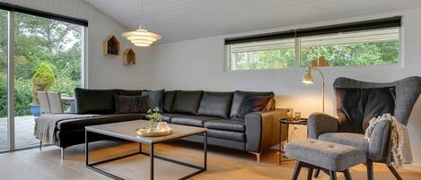 A modern living room with a sofa, a coffee table, and a chair. There are windows with blinds, a lamp, and a view of greenery outside.