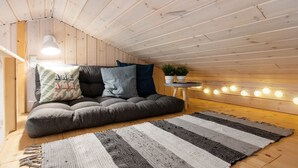 Een gezellig loft slaapkamer met een schuine kap, een grijze slaapbank met kussens, een gestreept vloerkleed en een klein bijzettafeltje met een plant.