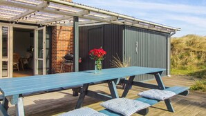 Een overdekt terras met een blauwe tafel en banken, een barbecue en een vaas met rode bloemen.