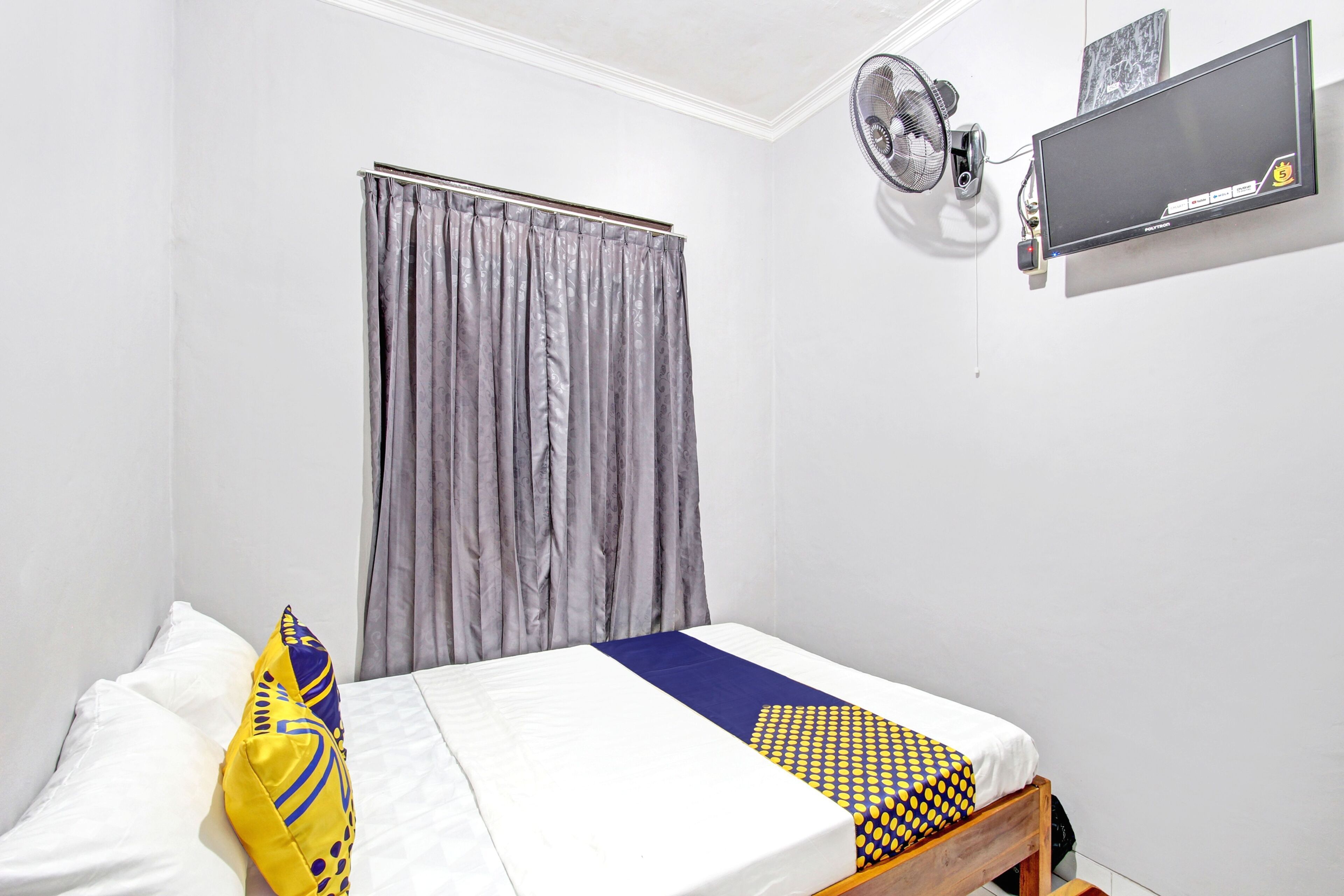 Une chambre à coucher avec un lit, une fenêtre avec des rideaux, un ventilateur fixé au mur et un téléviseur à écran plat.