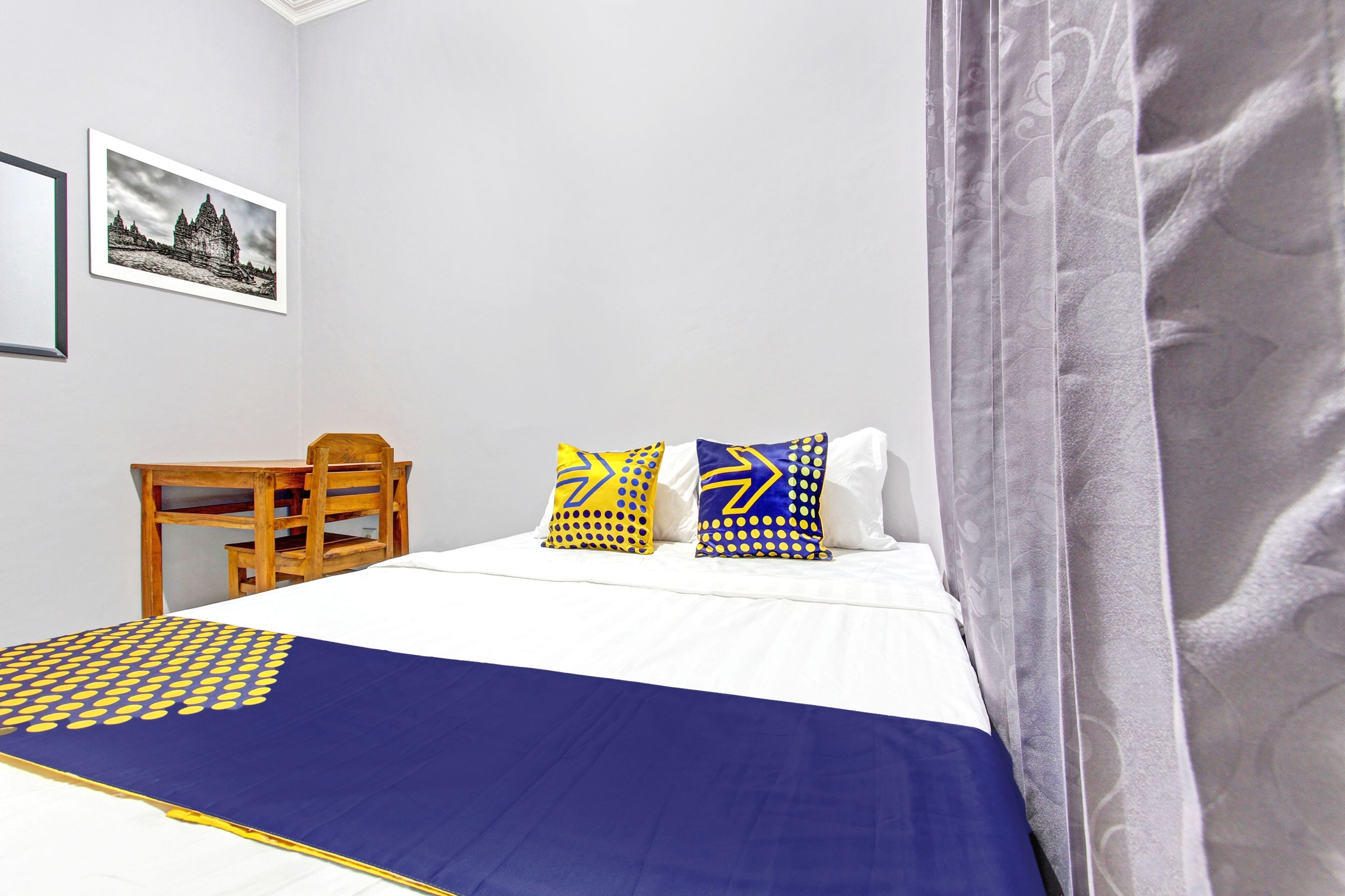 A hotel room with a bed, two pillows with geometric patterns, a chair, a framed picture on the wall, and a curtain.