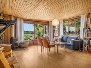 A cozy living room with a wooden ceiling, a fireplace, a sofa, a bookshelf, and a dining area with chairs.