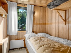 Habitación de cabaña de madera con cama, cortinas blancas, ventana con vista a árboles y una mesita de noche pequeña.