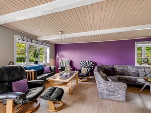 En moderne stue med lilla væg, trægulv og forskellige siddepladser, herunder en sofa, stole og en gyngestol.
