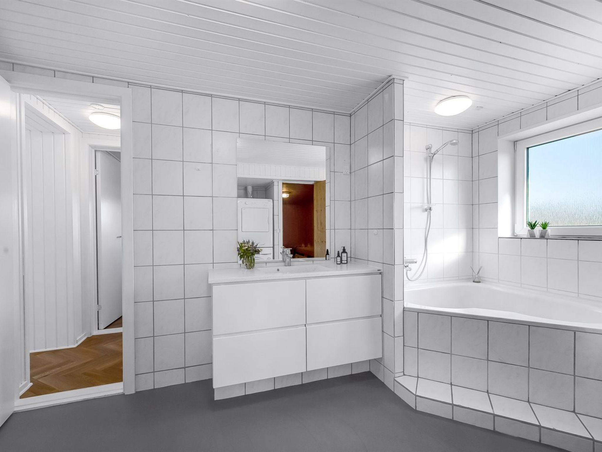 Ein modernes Badezimmer mit weißen Fliesen, einem großen Spiegel, einer Duschkabine und einem Fenster mit Pflanzen.