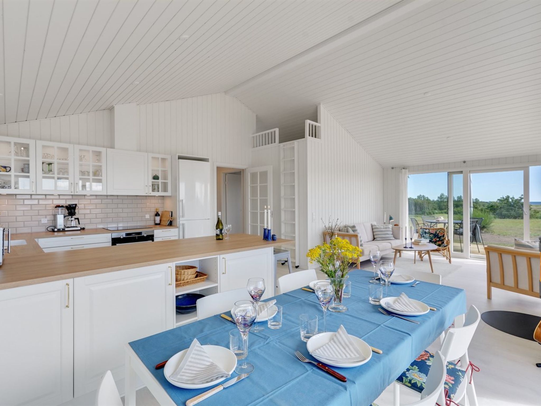 Una cucina moderna con mobili bianchi e un piano di lavoro in legno, un'area pranzo con tovaglia blu e tavolo apparecchiato, e vista all'esterno attraverso ampie porte scorrevoli in vetro.