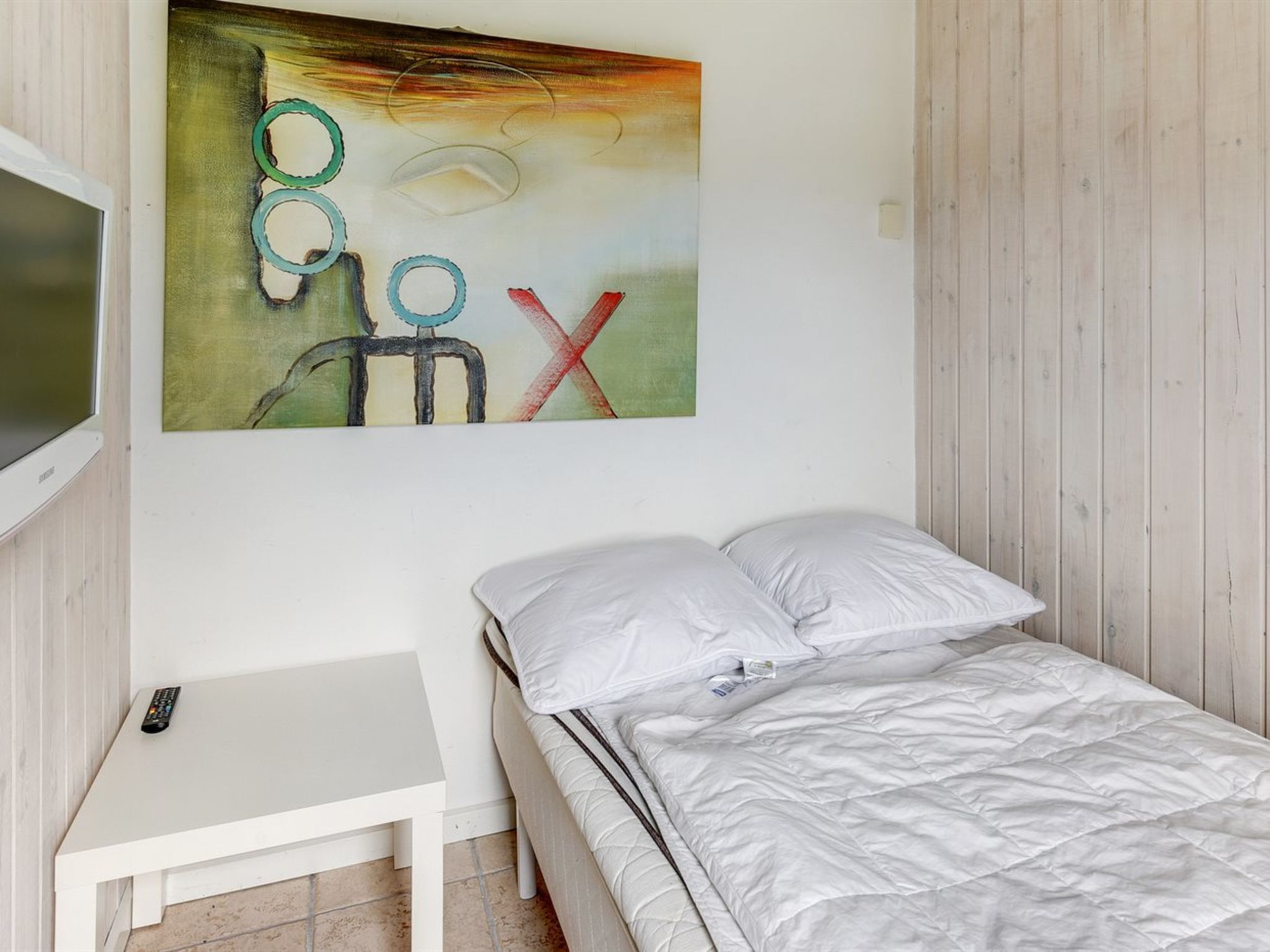 Ein Schlafzimmer mit einem Bett, einem kleinen Nachttisch, einem Fernseher und einem Wandbild.