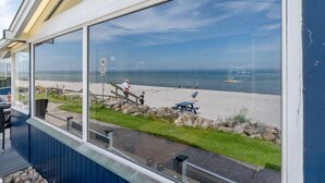 Vista desde una ventana con vista a una playa, cielo despejado, una terraza de madera y una mesa de picnic.