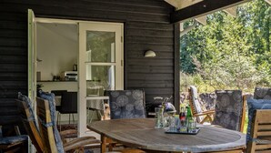An outdoor patio with a wooden table and chairs, a glass door leading to a kitchen area, and a view of greenery. - "Kringfod" - 590m from the sea by Interhome (Hals)